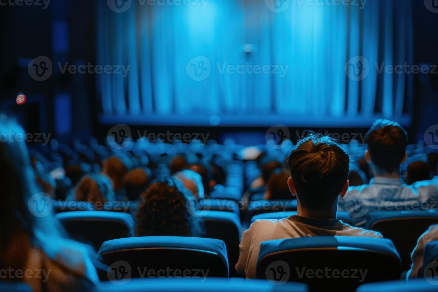 An audience sits in a darkened theater, captivated by a performance on stage, with soft blue lighting highlighting their silhouettes. photo
