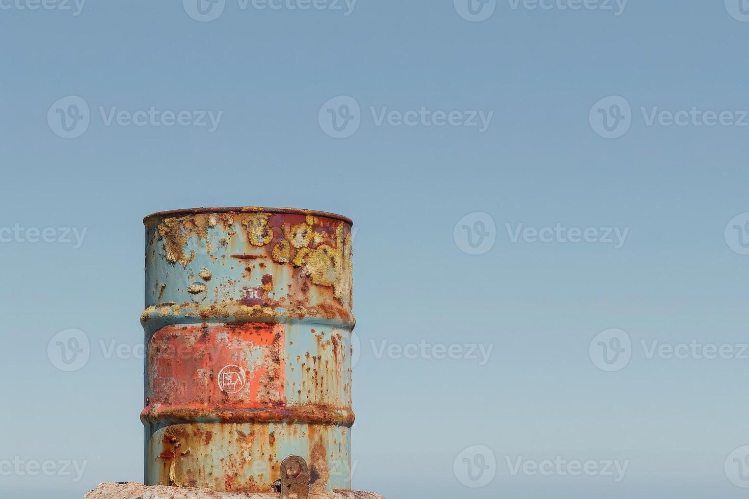 A weathered oil drum stands tall, showcasing vibrant colors and a distinct logo under a bright blue sky, evoking a sense of isolation and resilience in nature. photo