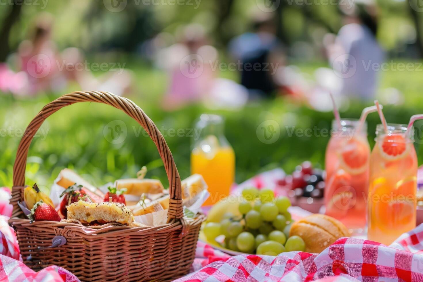 A picnic basket filled with sandwiches, fruit, and drinks sits on a red and white checkered blanket in a lush green park, while friends enjoy the sunny day. photo