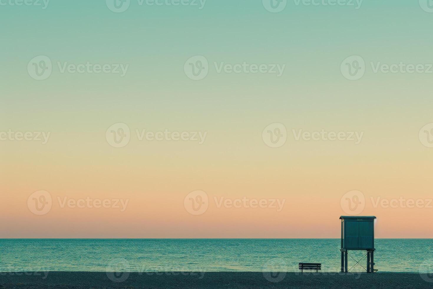 A solitary bench sits on the sandy shore overlooking a calm ocean, a small lifeguard stand silhouetted against the pastel sky of a peaceful sunrise. photo