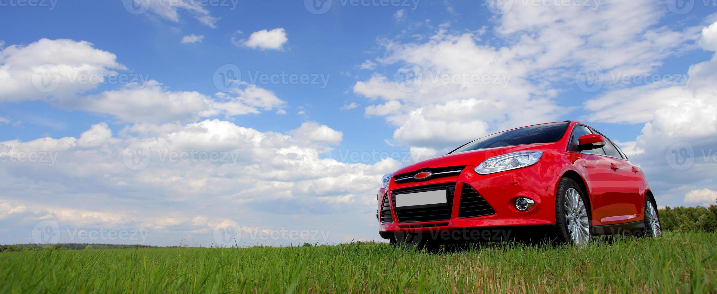 Red car on a blue sky background with white clouds. The car is standing in the field on the green grass. photo