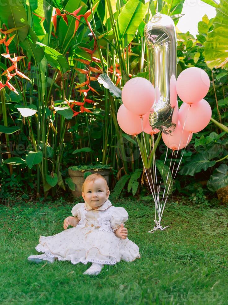 Birthday of cute baby girl with air balloons in summer garden. Happy child in dress. photo