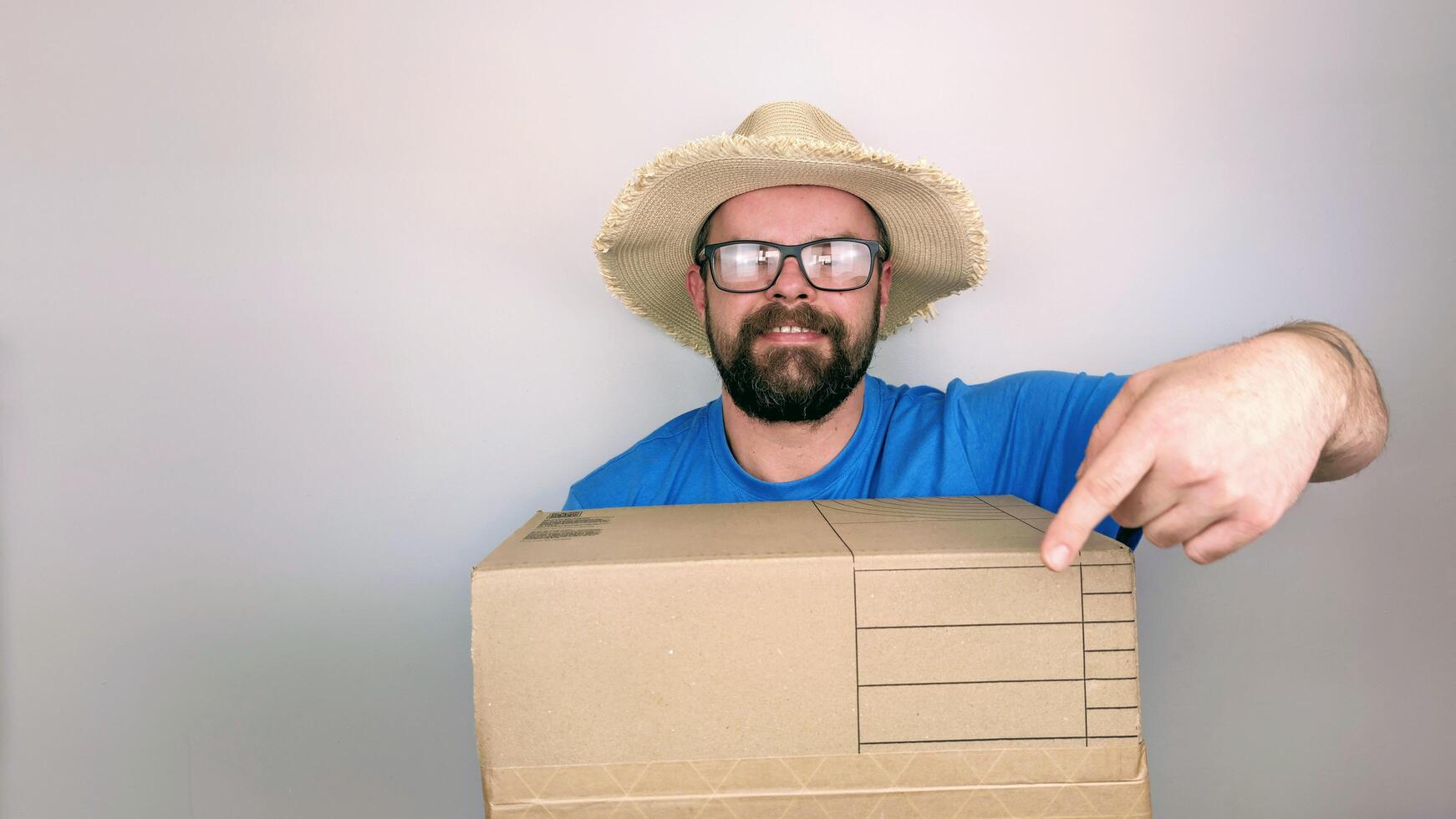 Portrait of bearded handsome delivery man in straw hat and blue shirt delivering box package, isolated on white background photo
