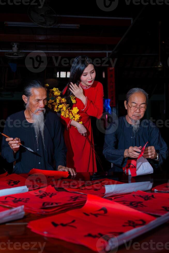 Vietnamese girl with red Ao Dai dress and old scholars writing calligraphy at lunar new year. Calligraphy is a popular tradition during Tet holiday. Text in photo mean Happy New Year and Peace.