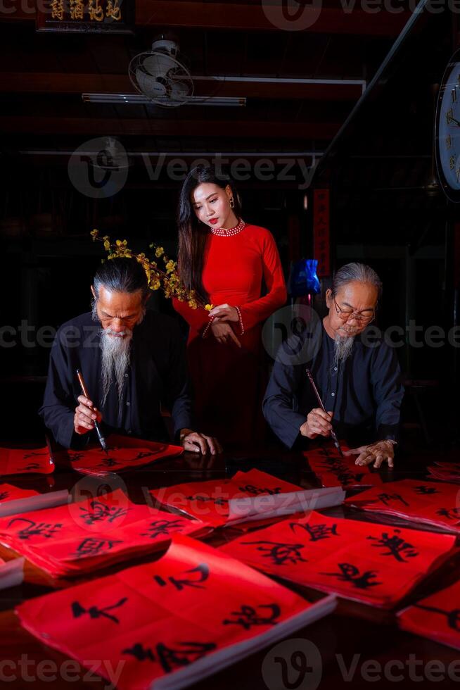 Vietnamese girl with red Ao Dai dress and old scholars writing calligraphy at lunar new year. Calligraphy is a popular tradition during Tet holiday. Text in photo mean Happy New Year and Peace.