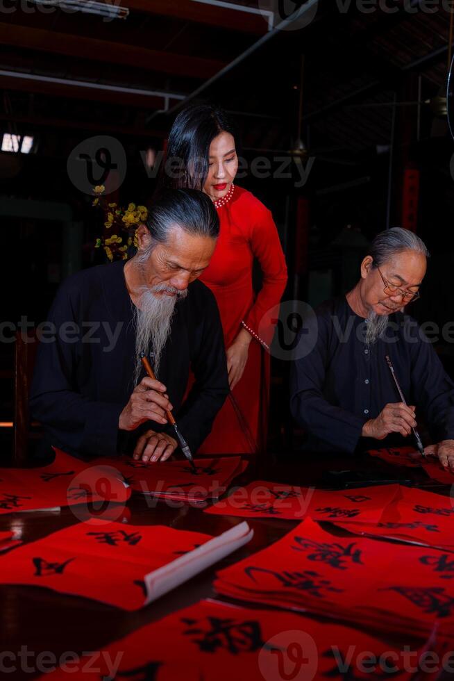 Vietnamese girl with red Ao Dai dress and old scholars writing calligraphy at lunar new year. Calligraphy is a popular tradition during Tet holiday. Text in photo mean Happy New Year and Peace.