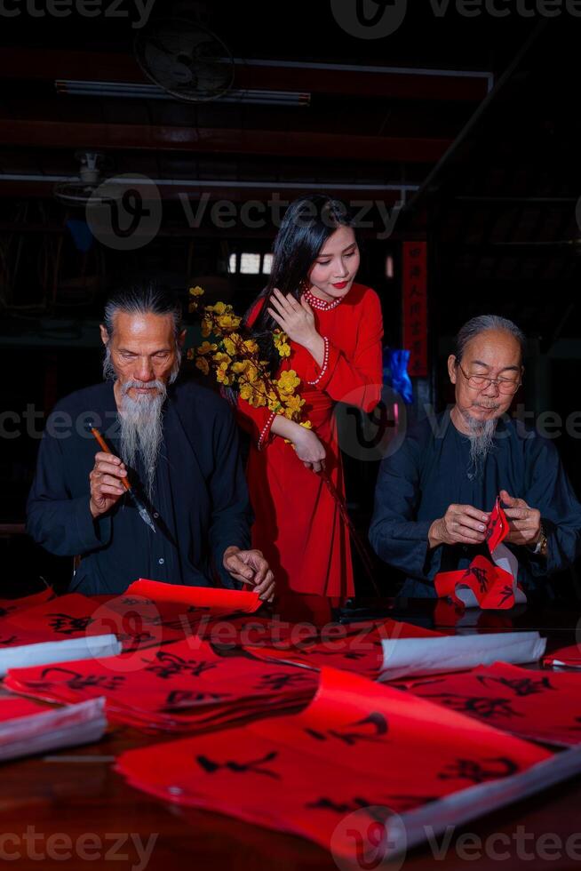Vietnamese girl with red Ao Dai dress and old scholars writing calligraphy at lunar new year. Calligraphy is a popular tradition during Tet holiday. Text in photo mean Happy New Year and Peace.