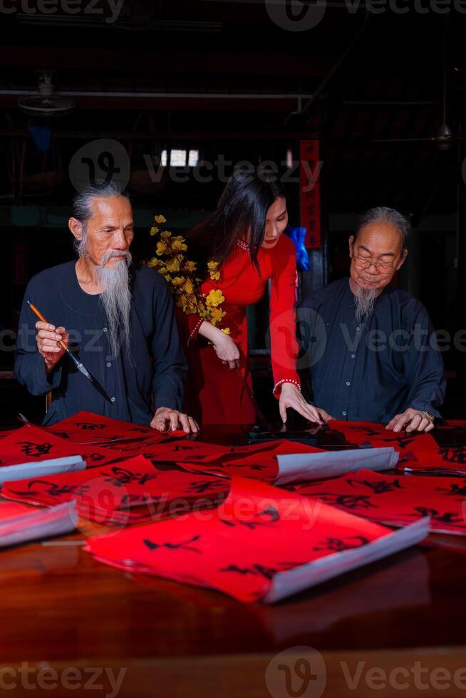Vietnamese girl with red Ao Dai dress and old scholars writing calligraphy at lunar new year. Calligraphy is a popular tradition during Tet holiday. Text in photo mean Happy New Year and Peace.