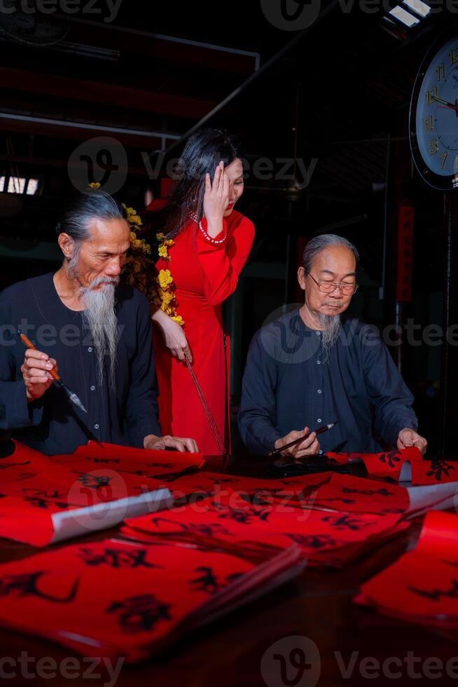 Vietnamese girl with red Ao Dai dress and old scholars writing calligraphy at lunar new year. Calligraphy is a popular tradition during Tet holiday. Text in photo mean Happy New Year and Peace.