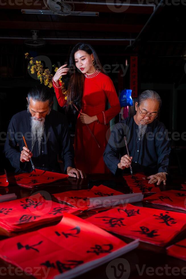 Vietnamese girl with red Ao Dai dress and old scholars writing calligraphy at lunar new year. Calligraphy is a popular tradition during Tet holiday. Text in photo mean Happy New Year and Peace.