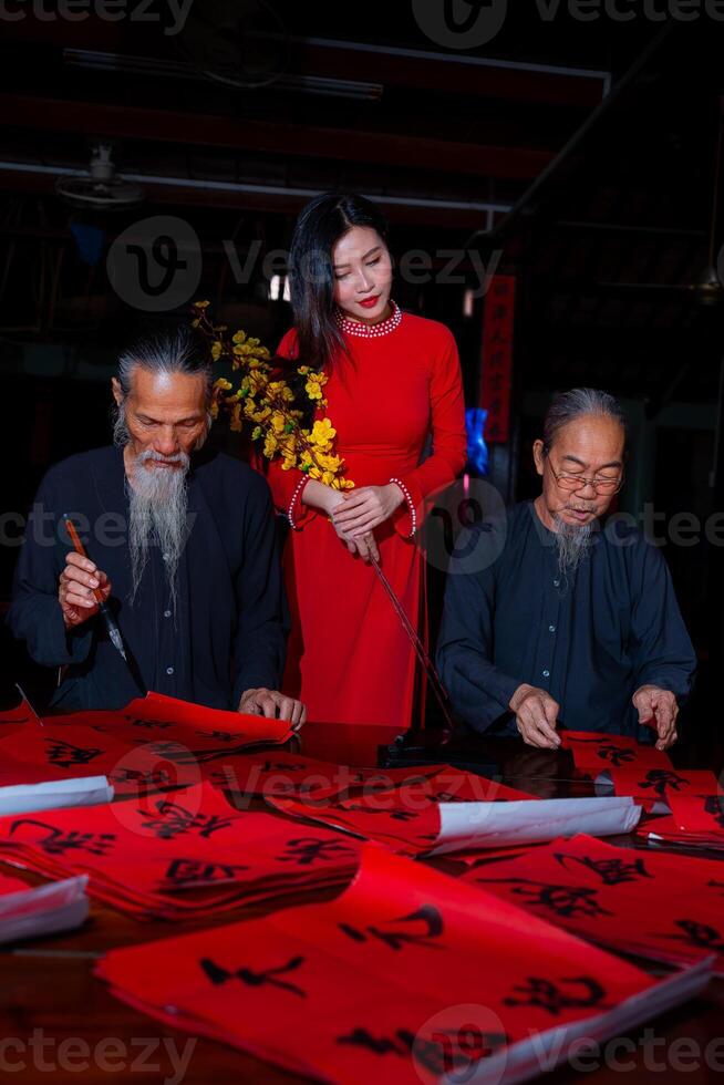 Vietnamese girl with red Ao Dai dress and old scholars writing calligraphy at lunar new year. Calligraphy is a popular tradition during Tet holiday. Text in photo mean Happy New Year and Peace.
