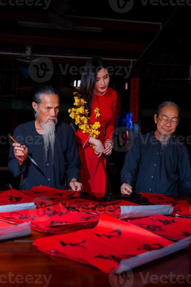 Vietnamese girl with red Ao Dai dress and old scholars writing calligraphy at lunar new year. Calligraphy is a popular tradition during Tet holiday. Text in photo mean Happy New Year and Peace.