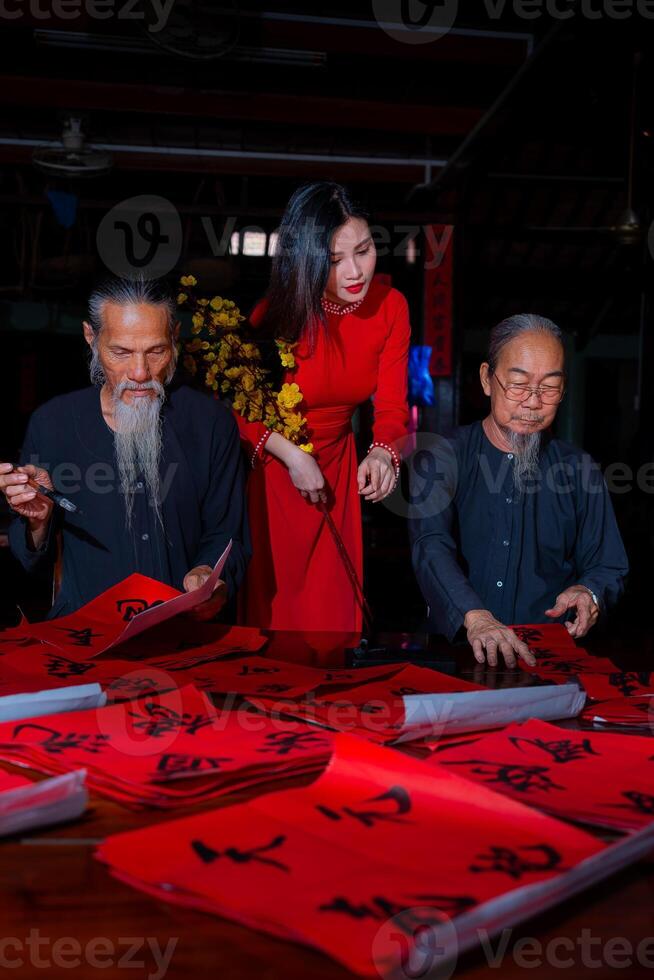 Vietnamese girl with red Ao Dai dress and old scholars writing calligraphy at lunar new year. Calligraphy is a popular tradition during Tet holiday. Text in photo mean Happy New Year and Peace.