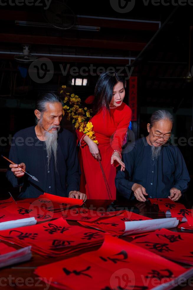 Vietnamese girl with red Ao Dai dress and old scholars writing calligraphy at lunar new year. Calligraphy is a popular tradition during Tet holiday. Text in photo mean Happy New Year and Peace.