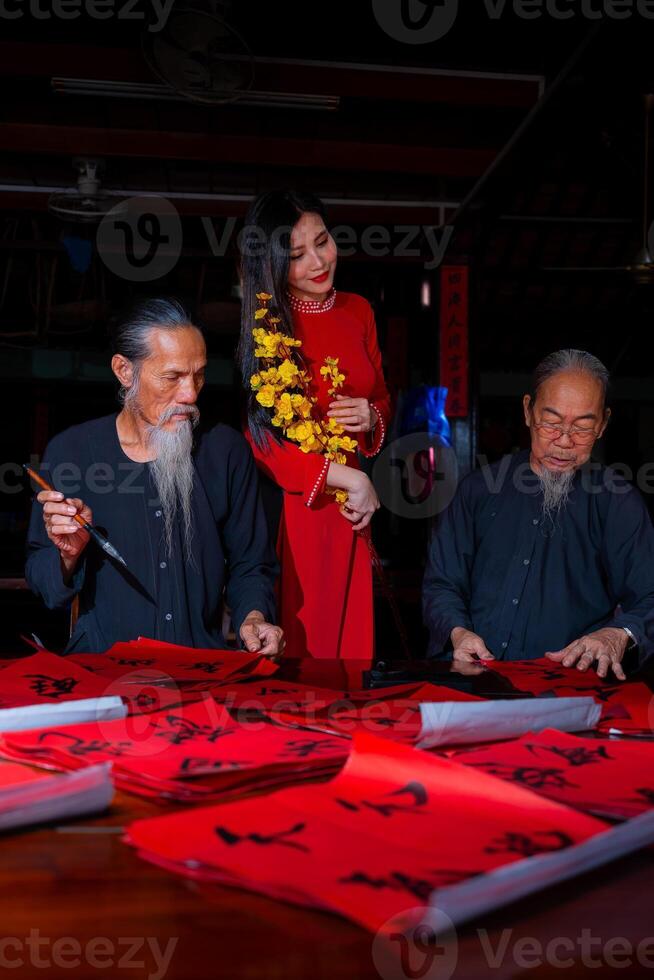Vietnamese girl with red Ao Dai dress and old scholars writing calligraphy at lunar new year. Calligraphy is a popular tradition during Tet holiday. Text in photo mean Happy New Year and Peace.