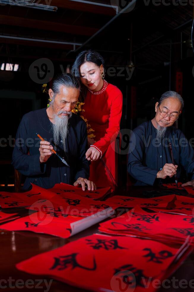 Vietnamese girl with red Ao Dai dress and old scholars writing calligraphy at lunar new year. Calligraphy is a popular tradition during Tet holiday. Text in photo mean Happy New Year and Peace.