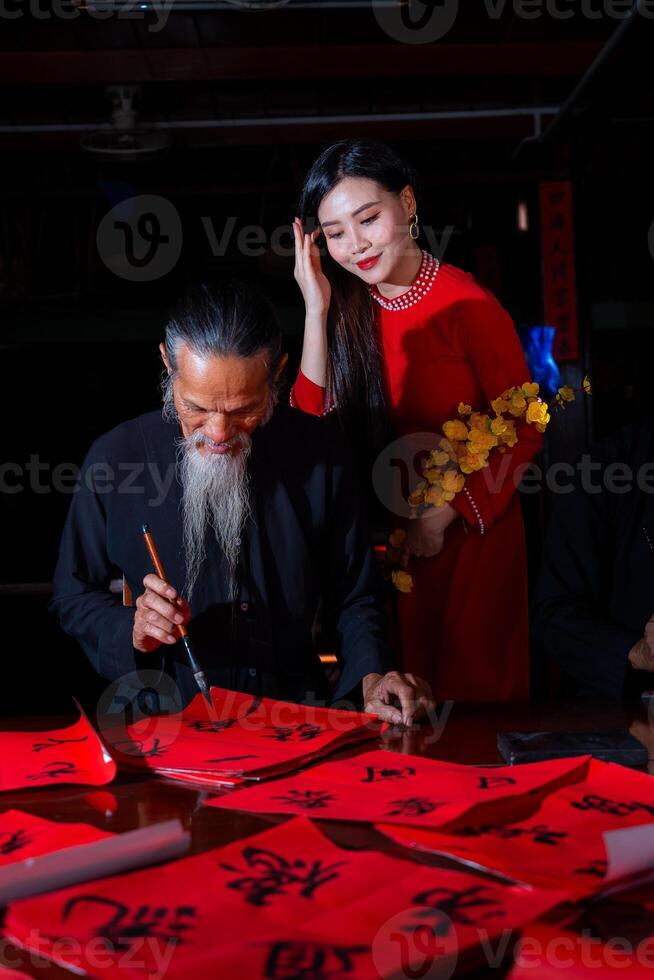 Vietnamese girl with red Ao Dai dress and old scholars writing calligraphy at lunar new year. Calligraphy is a popular tradition during Tet holiday. Text in photo mean Happy New Year and Peace.