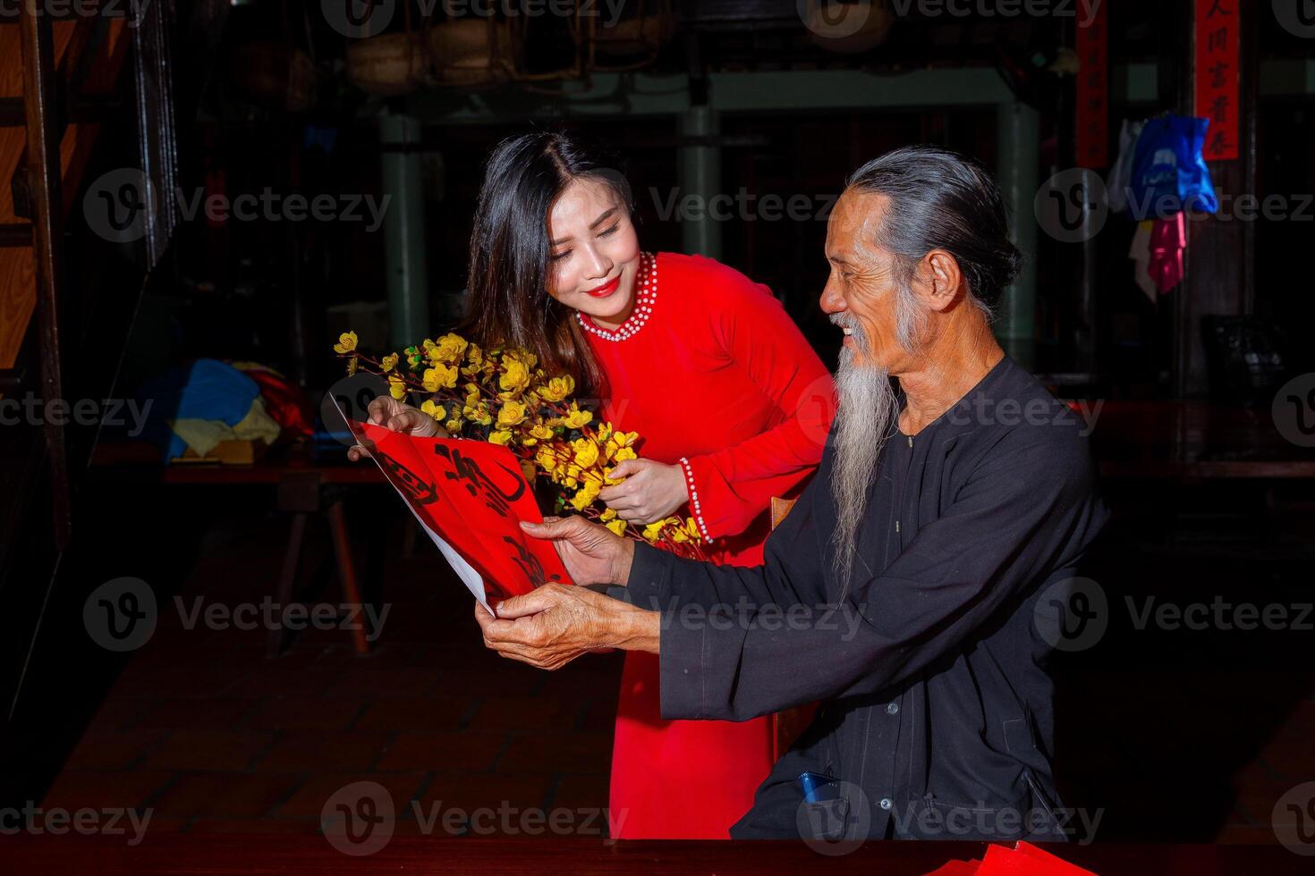 Vietnamese girl with red Ao Dai dress and old scholars writing calligraphy at lunar new year. Calligraphy is a popular tradition during Tet holiday. Text in photo mean Happy New Year and Peace.