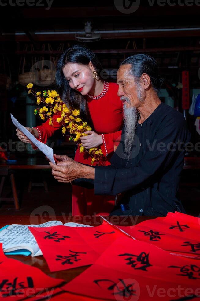 Vietnamese girl with red Ao Dai dress and old scholars writing calligraphy at lunar new year. Calligraphy is a popular tradition during Tet holiday. Text in photo mean Happy New Year and Peace.