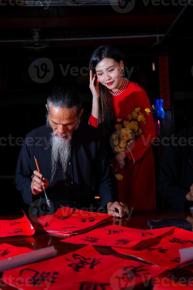 Vietnamese girl with red Ao Dai dress and old scholars writing calligraphy at lunar new year. Calligraphy is a popular tradition during Tet holiday. Text in photo mean Happy New Year and Peace.