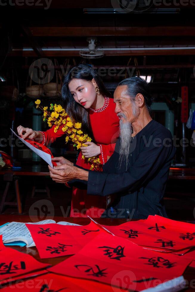 Vietnamese girl with red Ao Dai dress and old scholars writing calligraphy at lunar new year. Calligraphy is a popular tradition during Tet holiday. Text in photo mean Happy New Year and Peace.