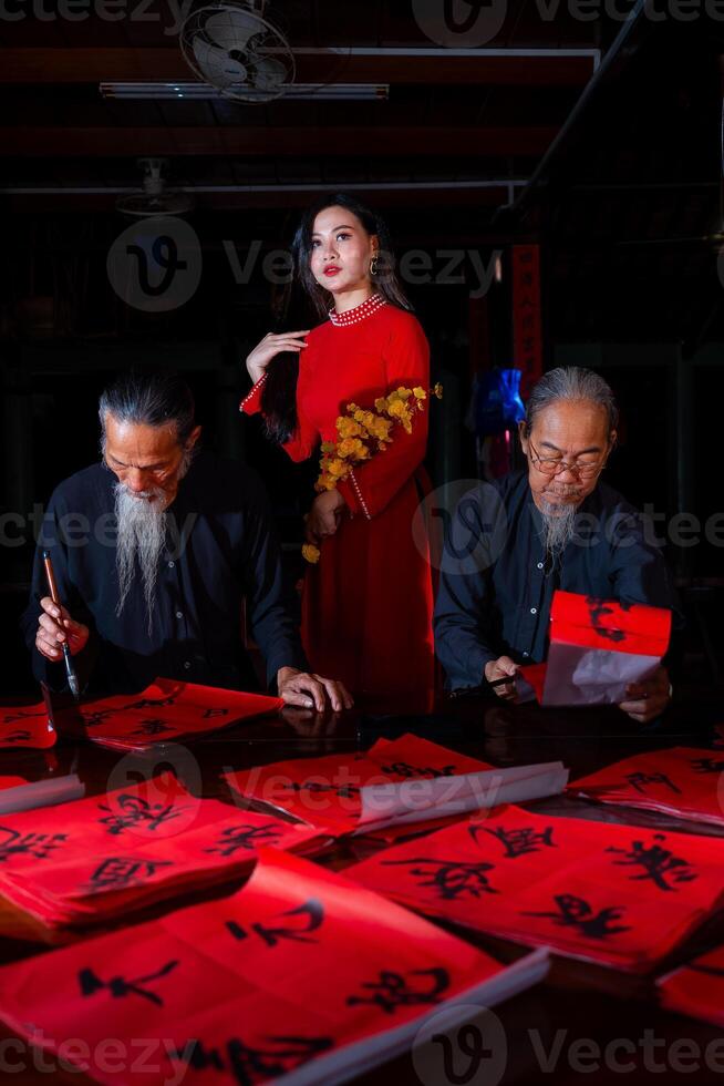 Vietnamese girl with red Ao Dai dress and old scholars writing calligraphy at lunar new year. Calligraphy is a popular tradition during Tet holiday. Text in photo mean Happy New Year and Peace.