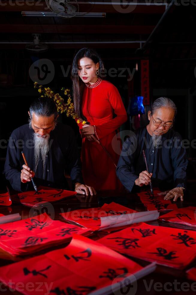 Vietnamese girl with red Ao Dai dress and old scholars writing calligraphy at lunar new year. Calligraphy is a popular tradition during Tet holiday. Text in photo mean Happy New Year and Peace.