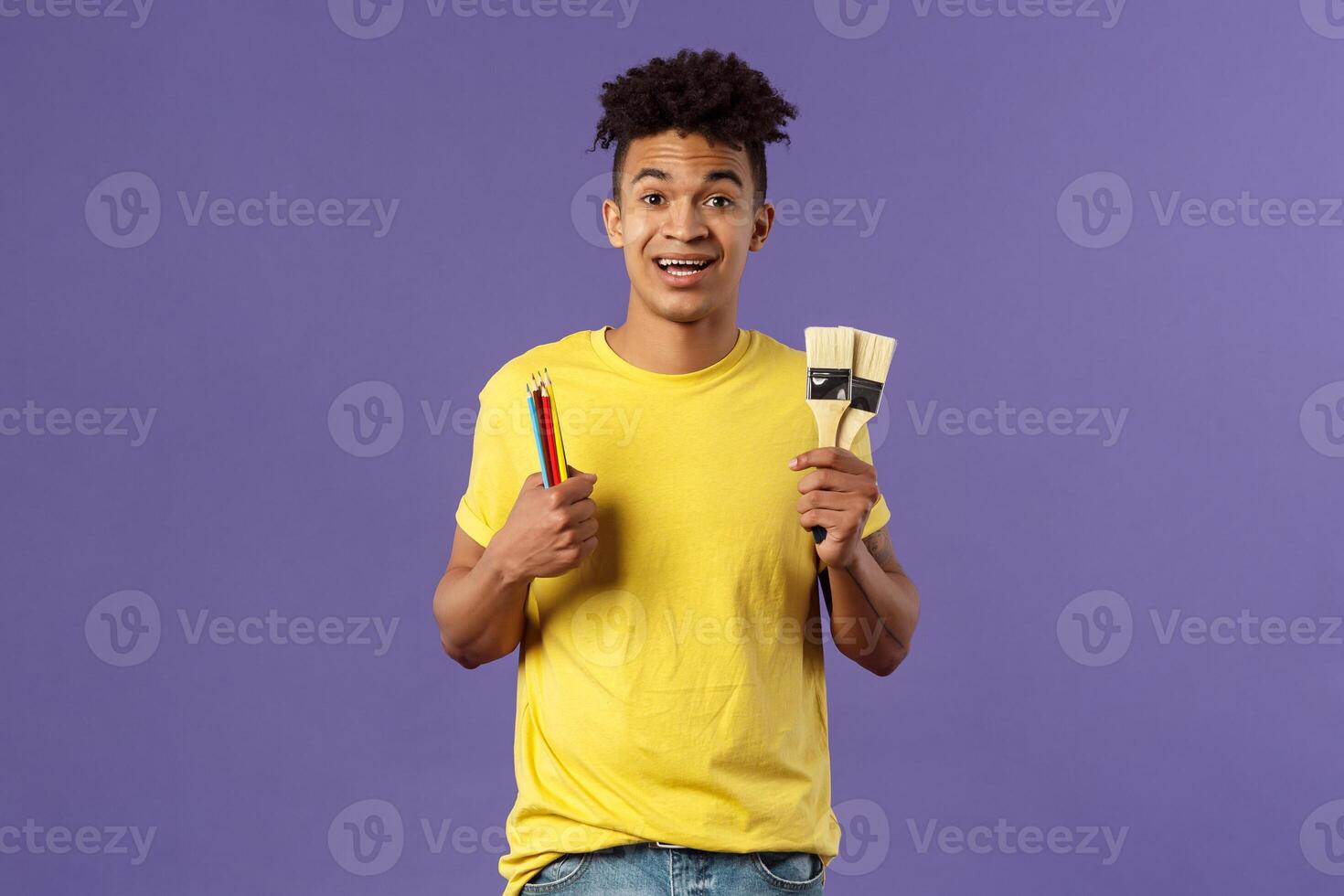 Portrait of upbeat young creative man, artist starting art courses, want to learn how to draw, holding colored pencils and brushes, standing enthusiastic over purple background photo