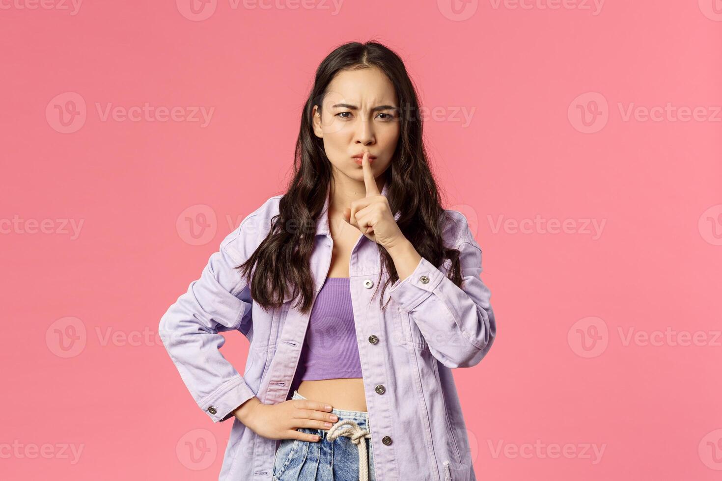 Shush dont speak. Serious-looking asian girl hush, frowning and looking upset, irritated with person speaking too loud, press index finger to lips, scolding, tell keep secret, pink background photo