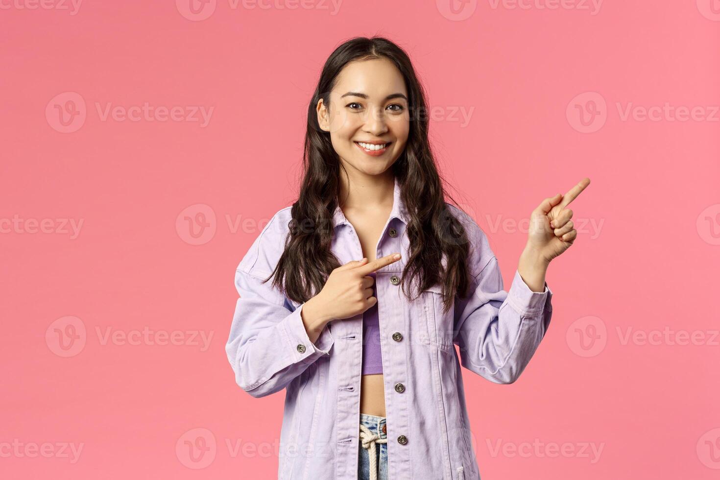 Portrait of cheerful asian girl, female student inviting people follow page, subscribe to page or visit link, pointing fingers right and smiling camera, standing happy over pink background photo