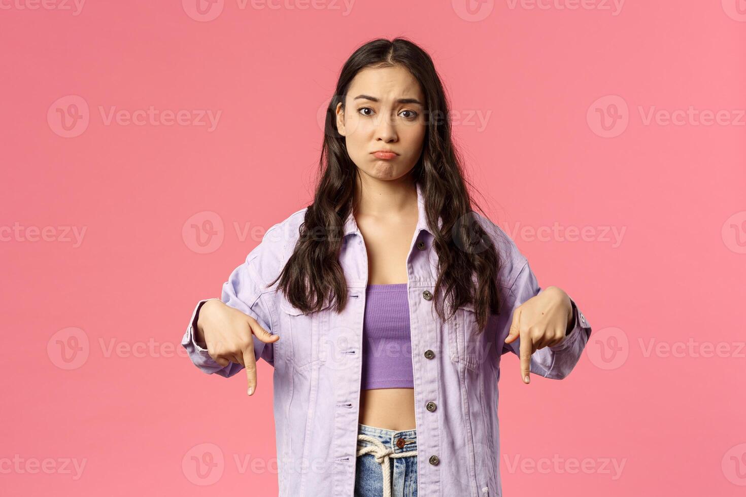 Displeased skeptical young asian girl pointing fingers down, pounting and making disappointed face, missed chance, lose in competition or see bad result, standing pink background unimpressed photo