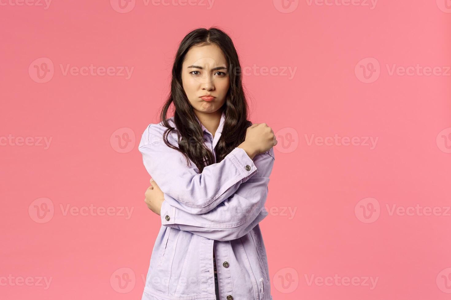 Portrait of silly and cute asian girl hugging herself and sulking, complaining on someone being rude and offensive to her, grimacing angry, waiting for apologies, standing pink background photo