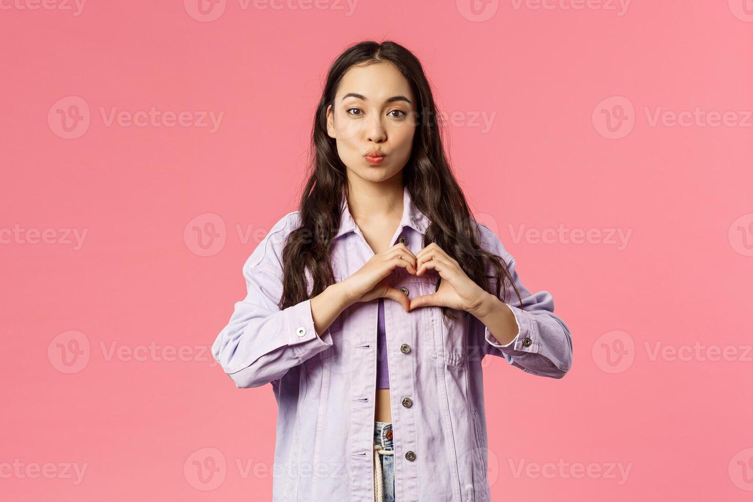 Portrait of romantic cute korean girl show heart gesture and fold lips, giving kiss, express sympathy, passion and romance, standing pink background in lovely stylish outfit photo