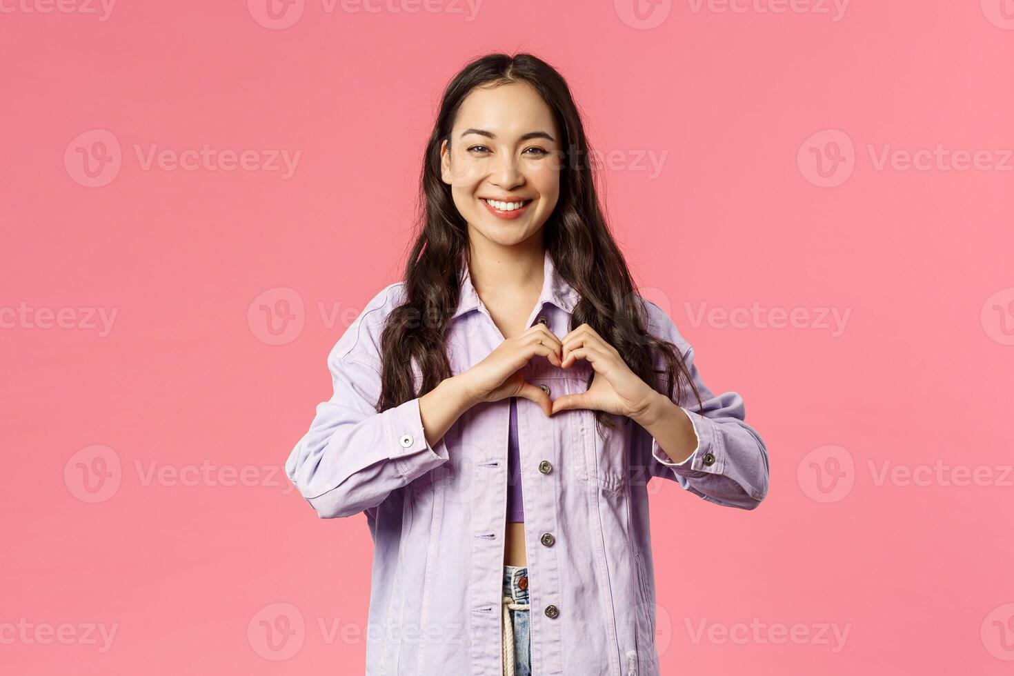 Portrait of cute, attractive asian girl in denim jacket, smiling as showing heart sign to confess love, being romantic, express sympathy or passionate feeling, falling for someone, pink background photo