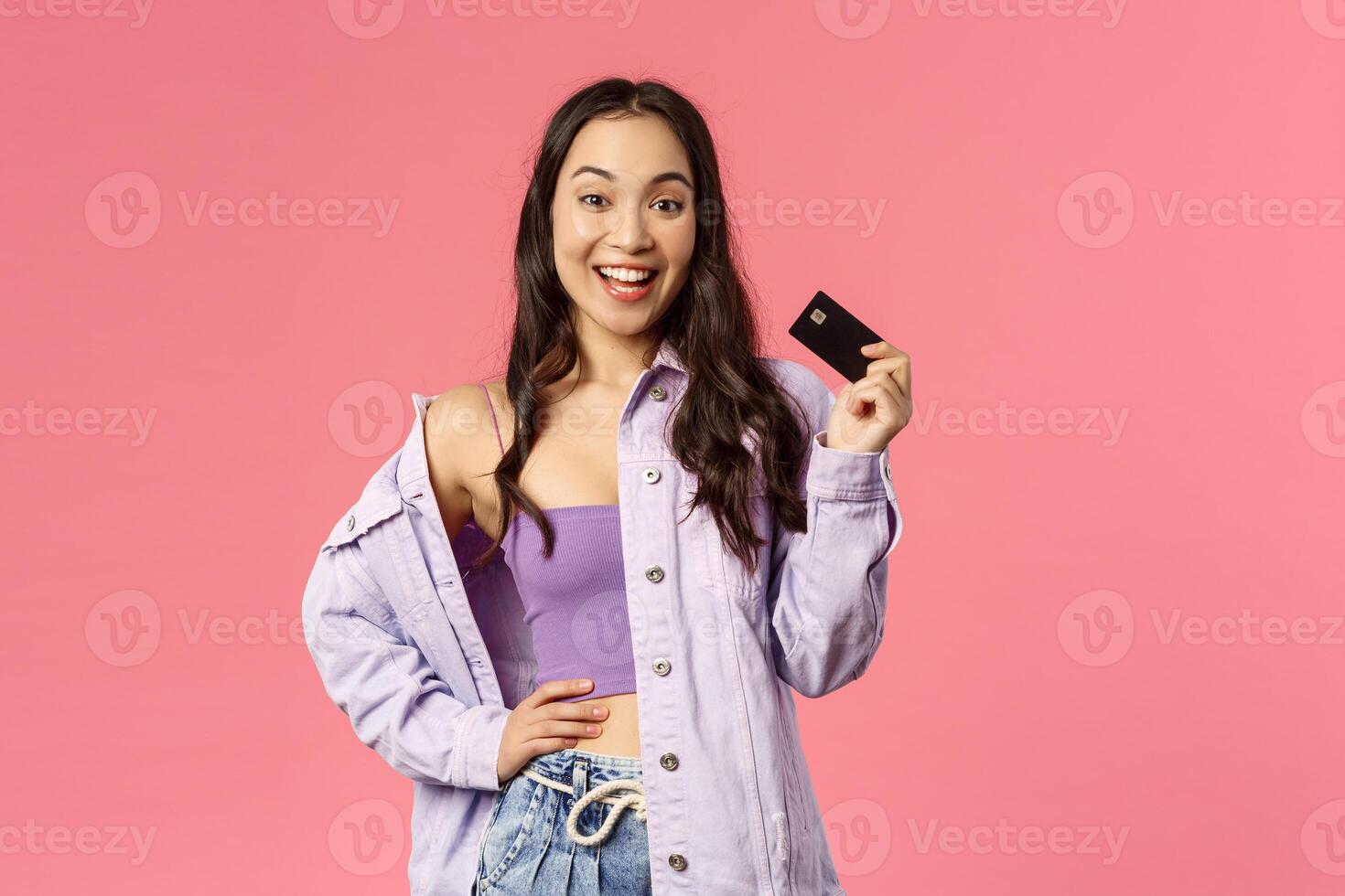 Look what Ive got. Portrait of stylish carefree girl, smiling and waving credit card as telling friends shes buying drinks tonight, ready for shopping, unbothered about money, pink background photo