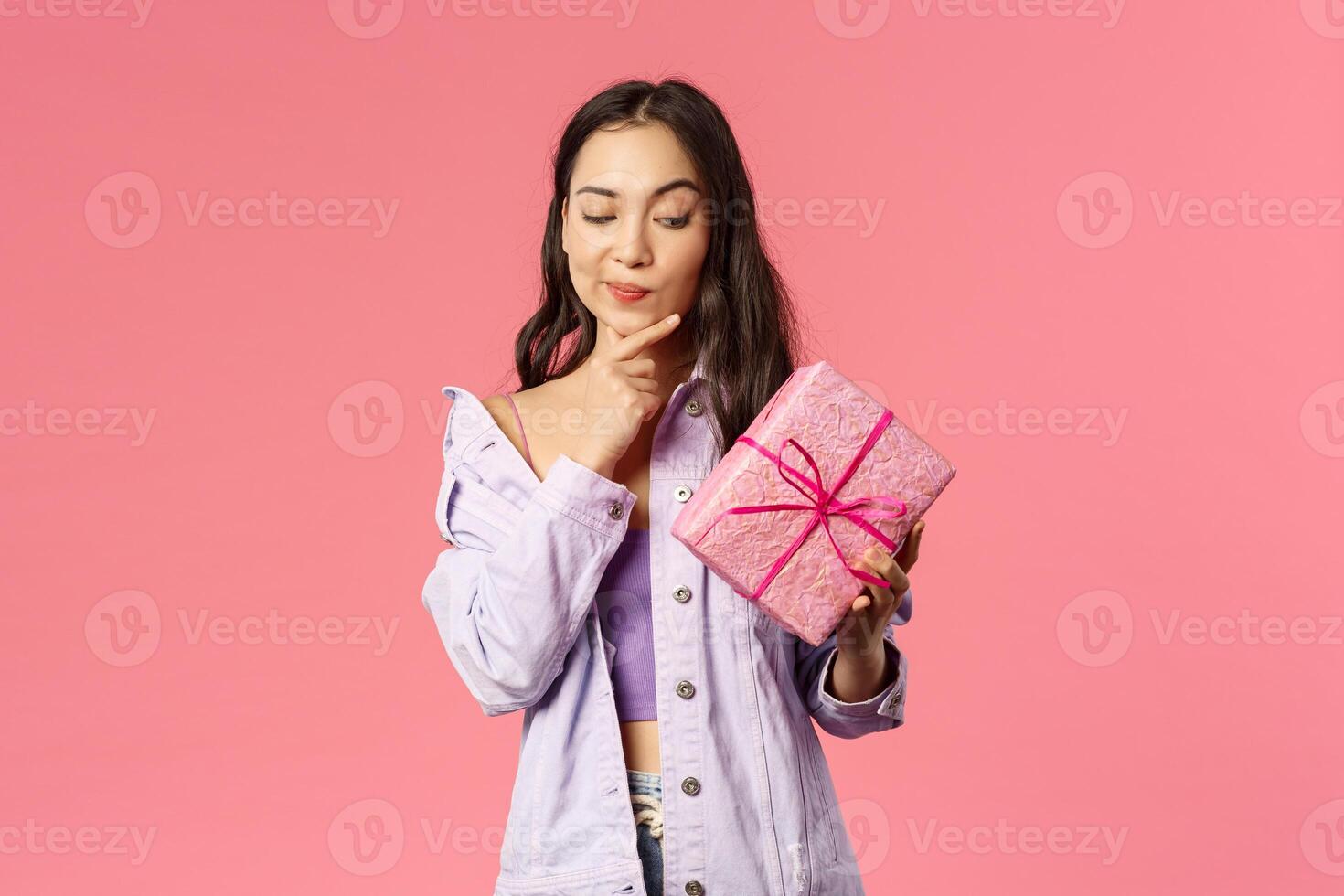 Portrait of curious enthusiastic young asian girl peeking at wrapped box, smirk and touching jaw as thinking about what inside of gift given by friend on birthday party, pink background photo