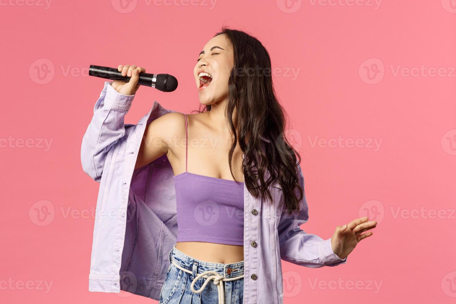 Portrait of excited good-looking, carefree asian girl singing with all her heart, put effort to reach that highest note, sing karaoke, close eyes and enjoying performing, standing pink background photo