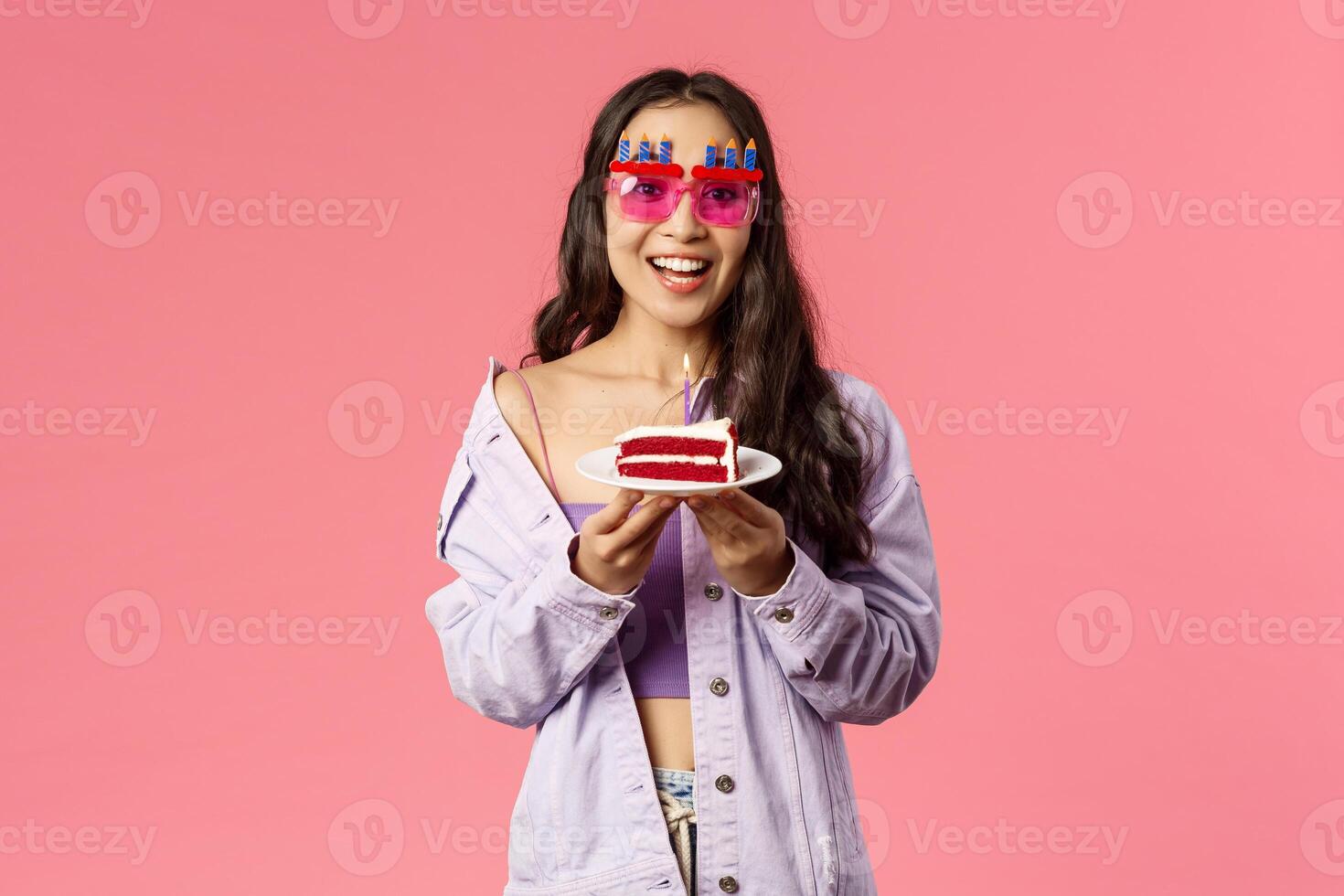 Celebration, holidays and people concept. Happy asian birthday girl smiling, wearing funny b-day glasses and eating cake, making wish on blowing candle, looking excited camera, pink background photo