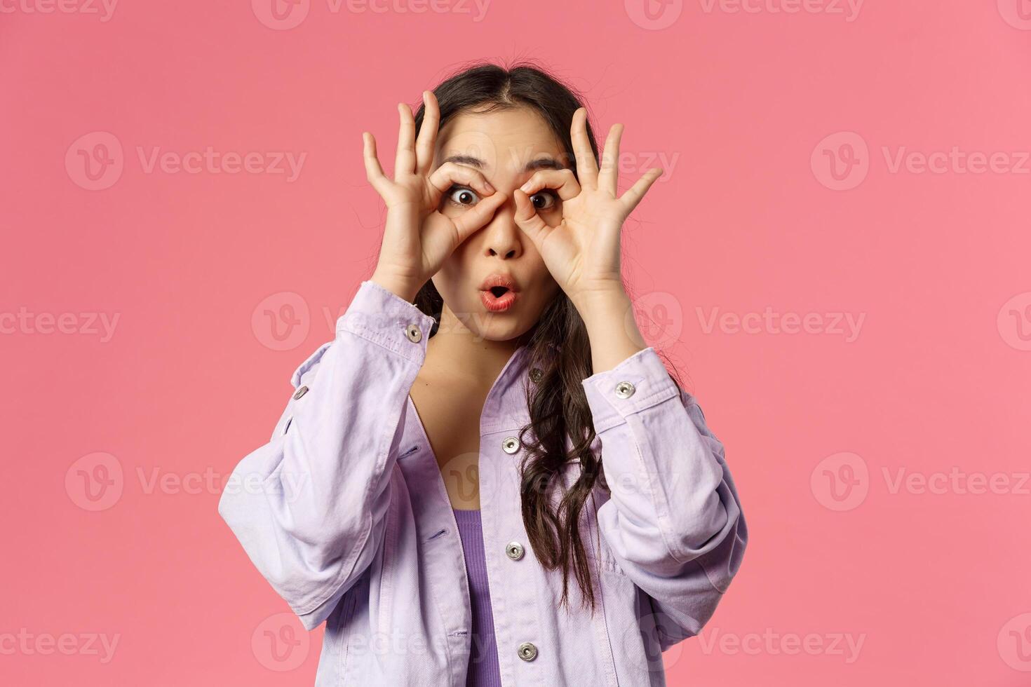 Close-up portrait of amused, speechless and impressed asian young girl seeing something curious and amazing, make glasses with fingers over eyes, fold lips say wow astonished, pink background photo