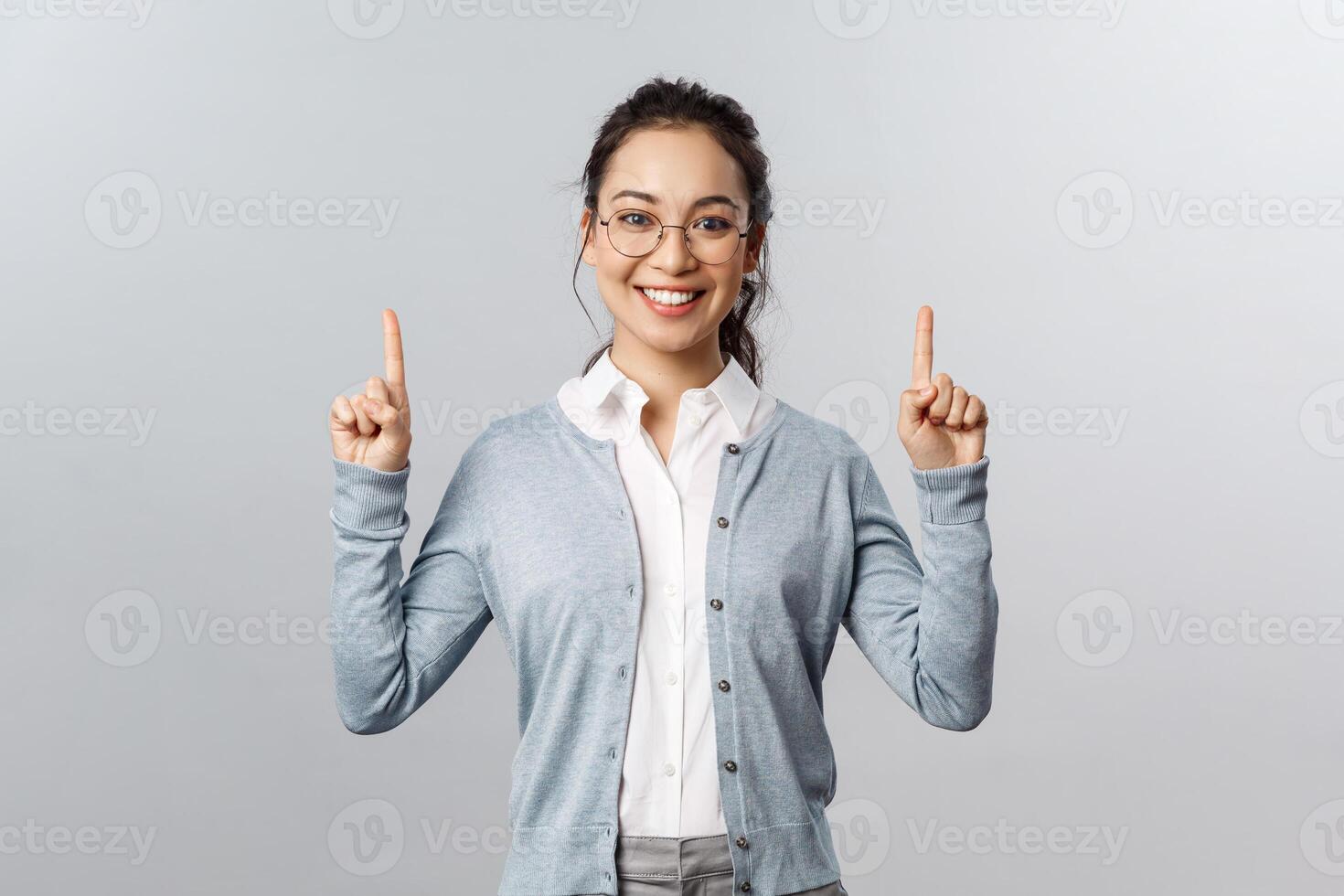 Friendly cheerful asian girl in glasses, showing product or company banner, pointing fingers up inviting check-up event news, info about advertisement, smiling adorable at camera, grey background photo