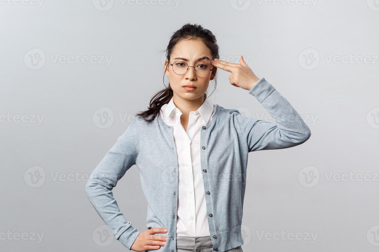Portrait of sick and tired asian woman hear something stupid, shoot herself with finger gun as a reaction to annoying bothering conversation, staring skeptical camera, standing grey background photo