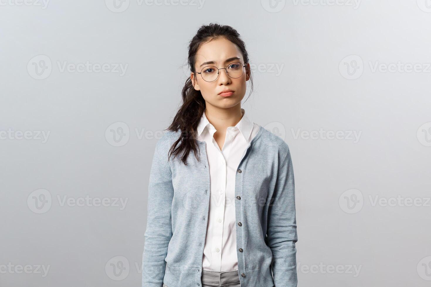 Gloomy and indifferent, lonely asian woman in glasses, feel uneasy and tired look camera drained, feel overworking and displeased, sighing with regret, standing upset over grey background photo