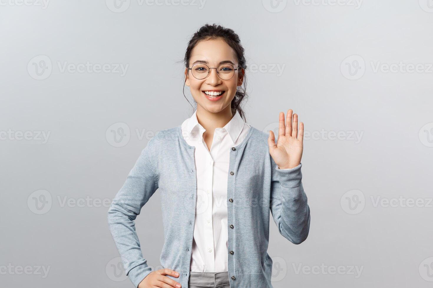 Lifestyle, people and emotions concept. Friendly enthusiastic asian woman in glasses, waving hand in hello, hi gesture, informal greeting of students during online lecture at app, smiling cheerful photo