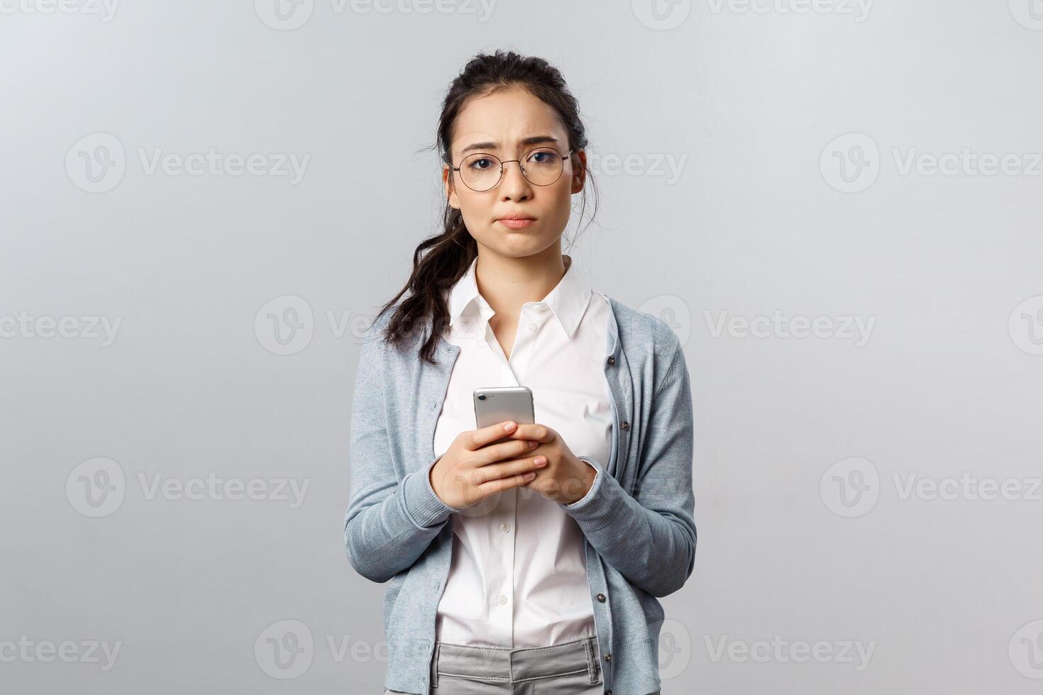 Office lifestyle, business and people concept. Troubled, uneasy young asian girl bothered and feel hesitant as sending risky text on dating app, look troubled camera, hold smartphone indecisive photo