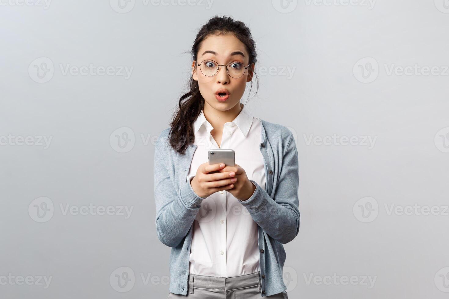 Office lifestyle, business and people concept. Amused and surprised asian girl impressed with awesome new app she uploaded on her smartphone, look camera astounded, use mobile phone application photo