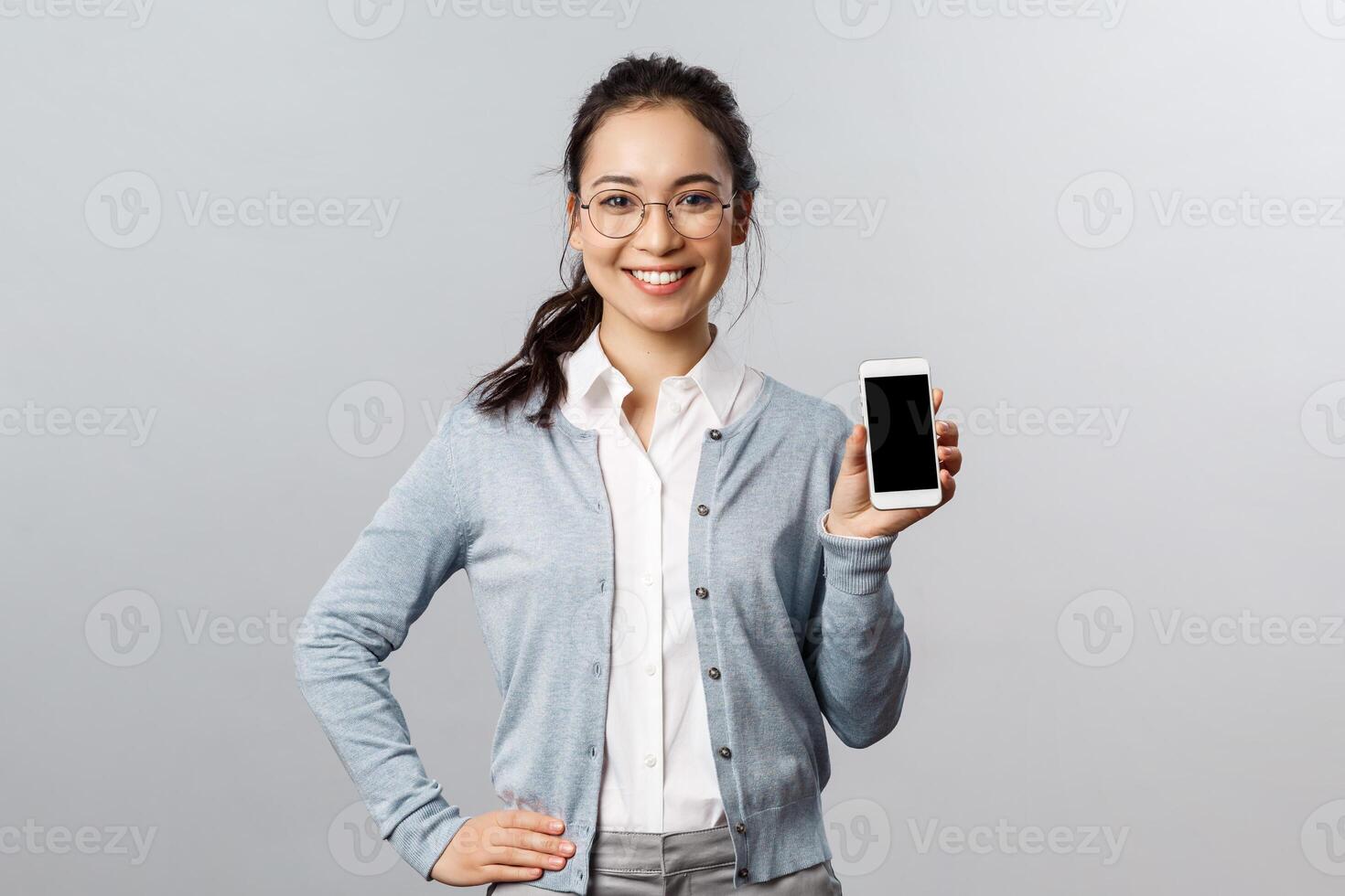 Technology, online and mobile lifestyle concept. Cheerful asian woman in glasses introduce new application, internet store link, showing smartphone display with pleased smile photo