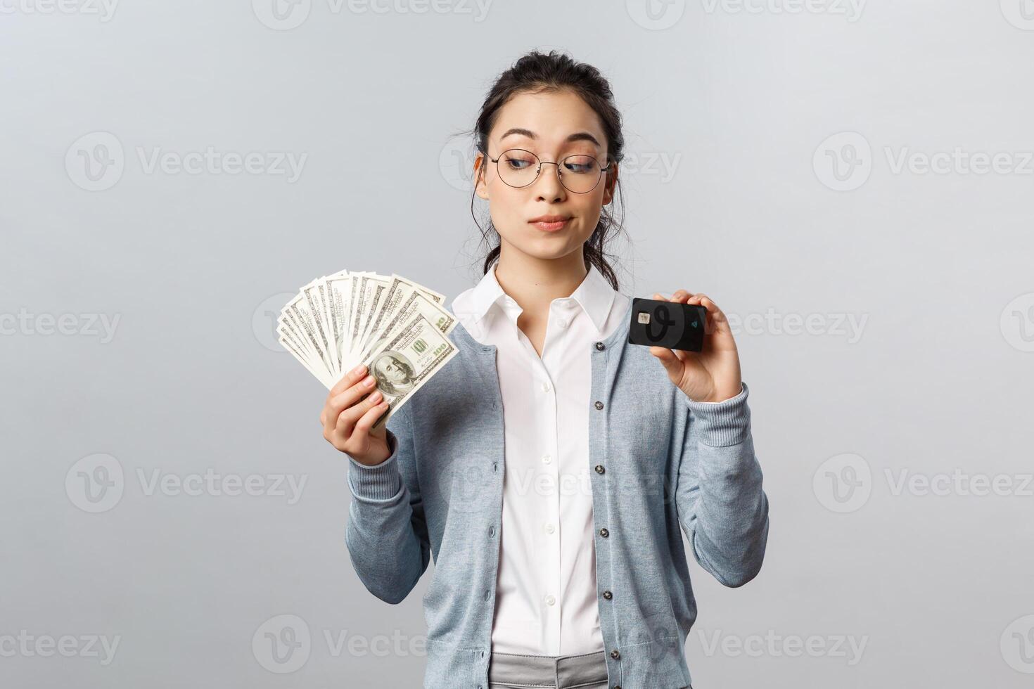 Business, finance and investment concept. Thoughtful indecisive cute asian girl look tempting at cash dollars, holding money and credit card, shopping, think where invest, grey background photo