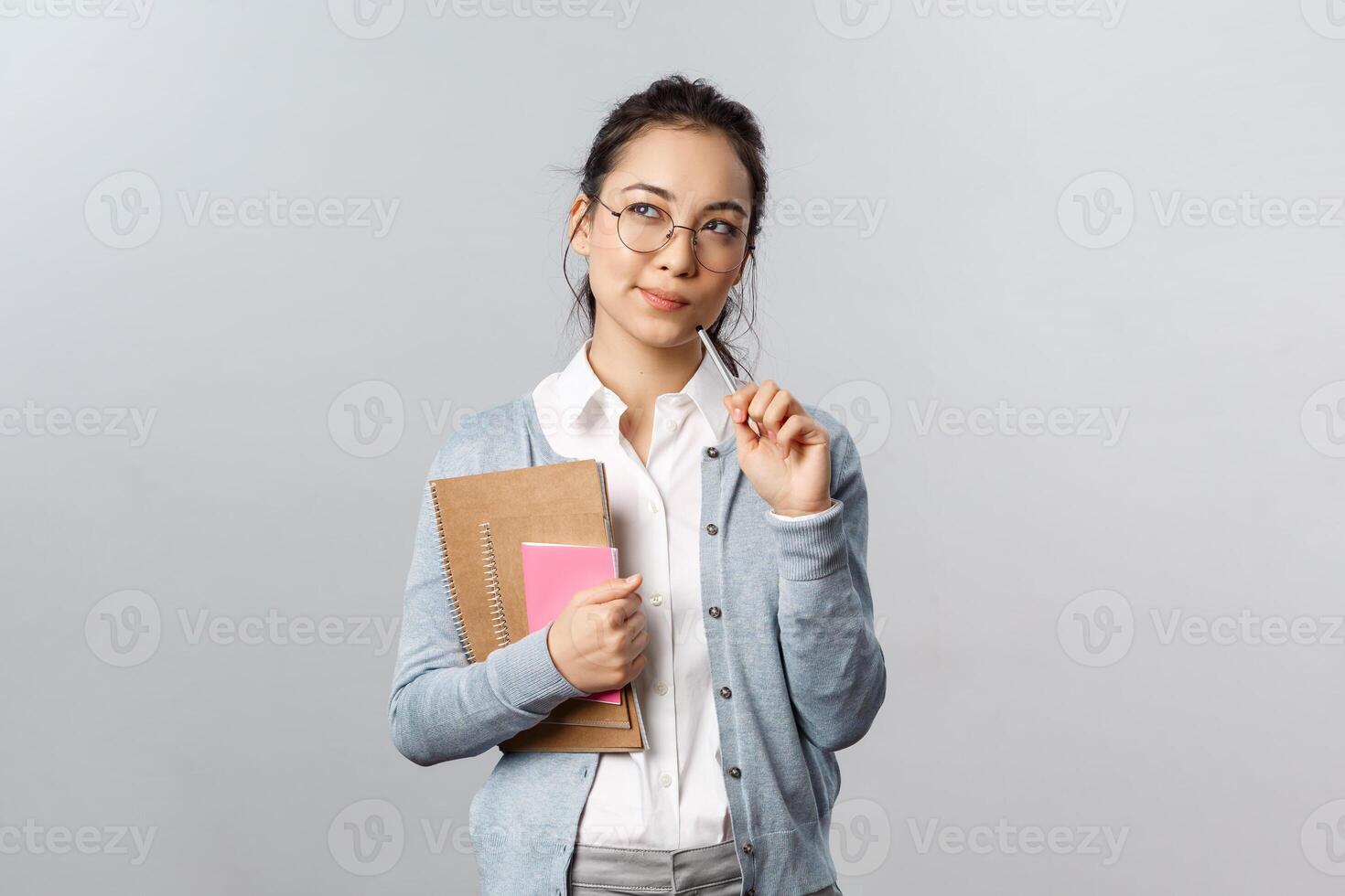 Education, teachers, university and schools concept. Thoughtful asian female student, solving puzzle, look up with intrigued, thinking face, bite pencil while pondering, hold notebooks photo