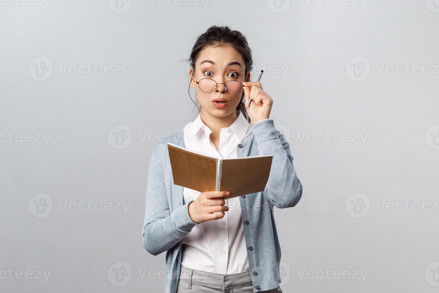 Education, teachers, university and schools concept. Surprised silly asian female student studying, holding notebooks and pen, fixing glasses look wondered at camera, hear question from student photo