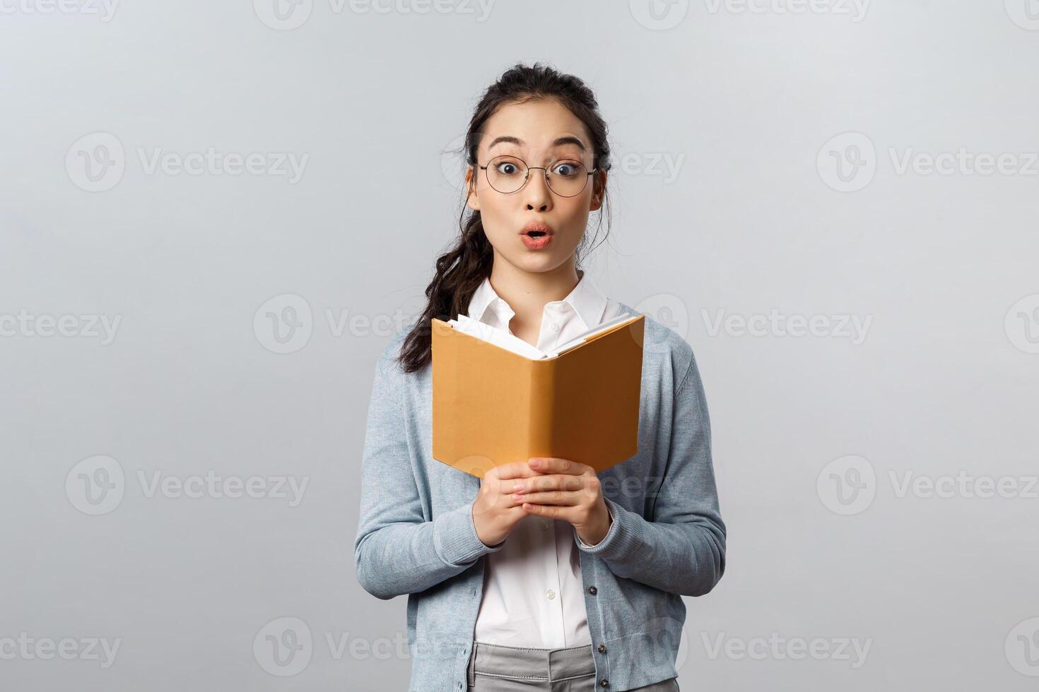 Education, teachers, university and schools concept. Amused and excited asian female tutor reading good homework of student in notebook, fold lips and look camera wondered, grey background photo